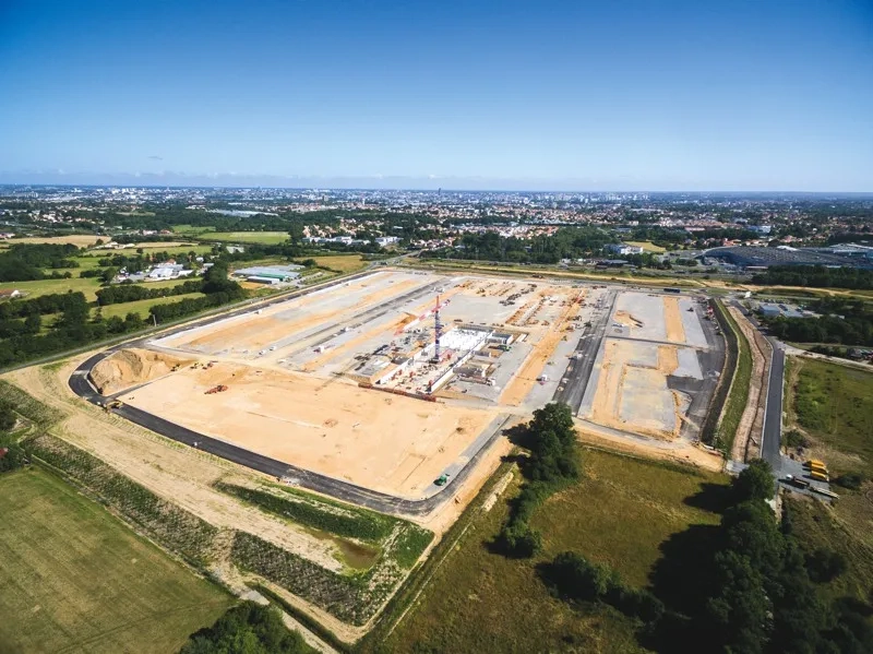 Deux vues aériennes du chantier, l’une en avril 2017 (où le traitement de plate-forme s’achève), l’autre deux mois plus tard, en juin 2017 (où la construction des premiers bâtiments commence). Deux vues aériennes du chantier, l’une en avril 2017 (où le traitement de plate-forme s’achève), l’autre deux mois plus tard, en juin 2017 (où la construction des premiers bâtiments commence).
