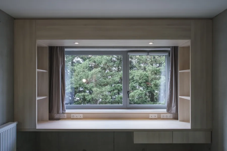 Aménagé devant la fenêtre, un meuble caisson en bois stratifié clair, de la largeur de la chambre, regroupe une table de travail, des espaces de rangement et la connectique.